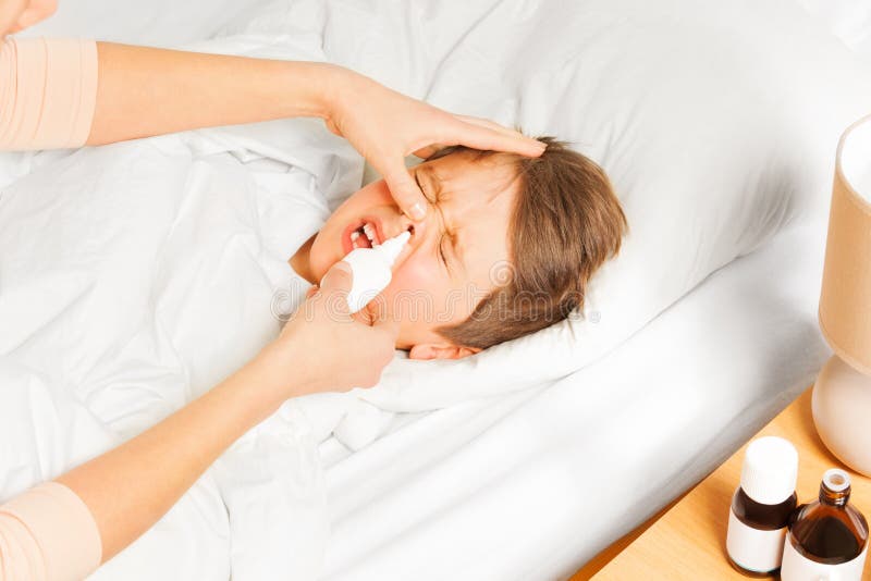 Sick five years old boy laying in bed and crying while mother spraying him nasal spray or drops in nose. Sick five years old boy laying in bed and crying while mother spraying him nasal spray or drops in nose