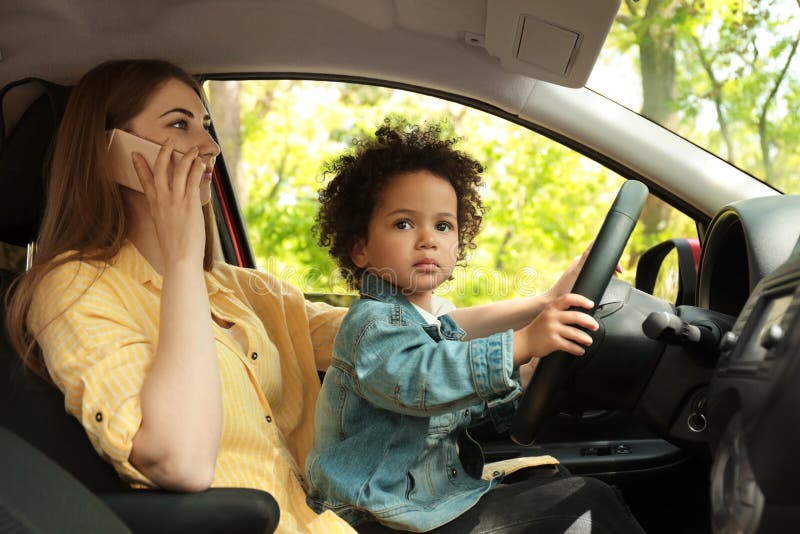 Madre con poca figlia sulle ginocchia che conducono automobile e che parlano dal telefono immagine stock Madre con poca figlia sulle ginocchia che conducono automobile e che parlano dal telefono immagine stock