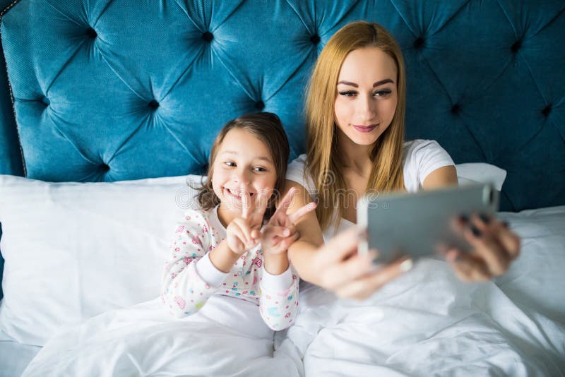 Madre e figlia che si trovano sul letto e che prendono autoritratto con lo smartphone Donna che prende selfie con una bambina in fotografia stock Madre e figlia che si trovano sul letto e che prendono autoritratto con lo smartphone Donna che prende selfie con una bambina in fotografia stock