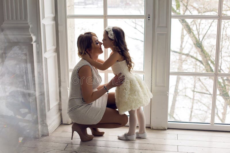 Mother and daughter three years sitting by the large window in white dresses before the new year. Mother and daughter three years sitting by the large window in white dresses before the new year