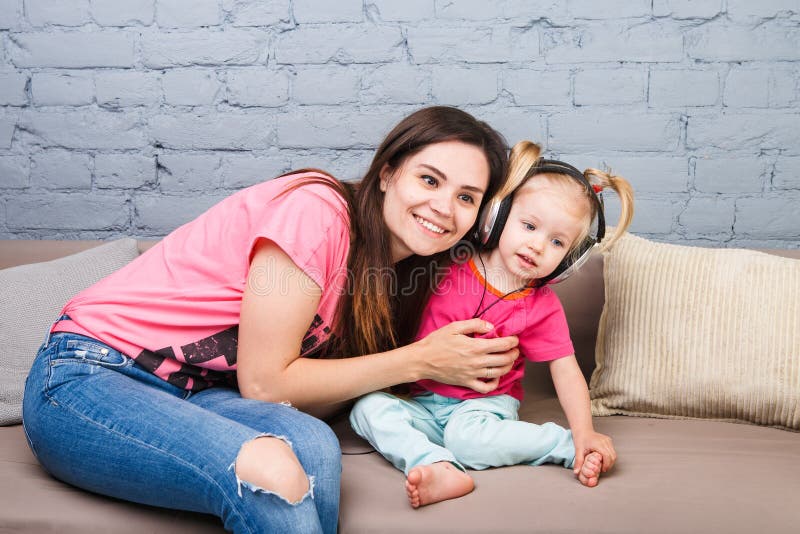 Madre e una figlia di due anni che ascoltano la musica con le cuffie sulla loro testa Sieda sullo strato nella stanza contro immagine stock libera da diritti Madre e una figlia di due anni che ascoltano la musica con le cuffie sulla loro testa Sieda sullo strato nella stanza contro immagine stock libera da diritti