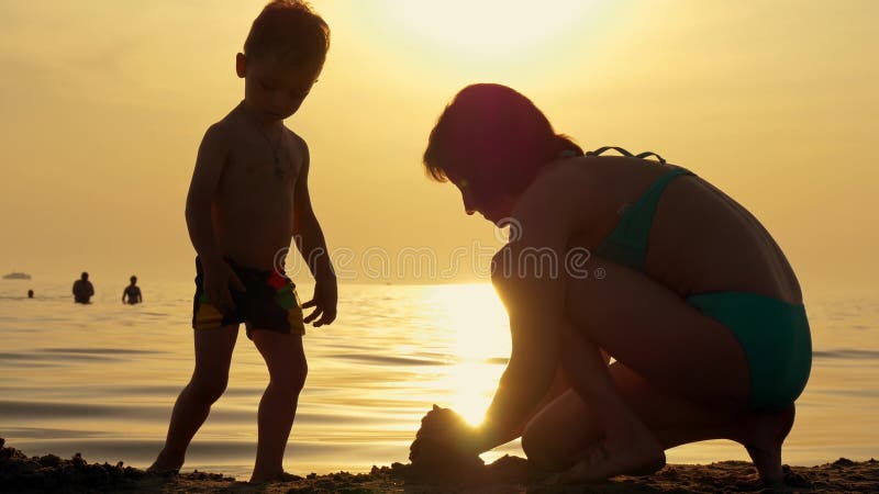 Madre feliz y niño que juegan con la arena en la playa contra puesta del sol