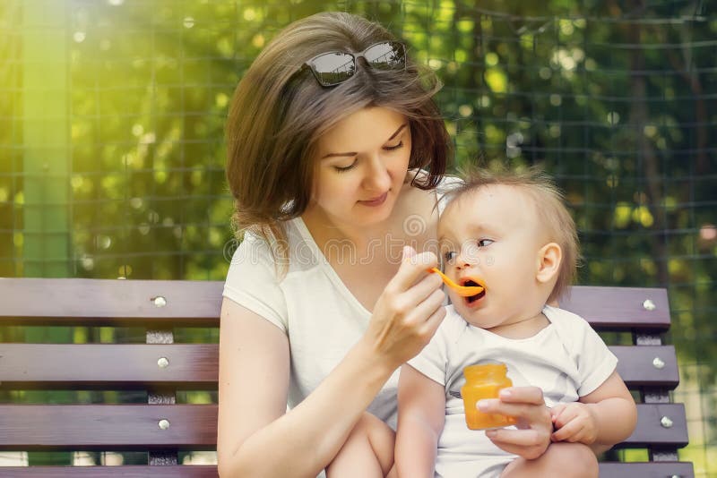 Madre que da a su niño infantil el puré de alimentación complementario de la calabaza en el día soleado al aire libre La mamá y e imágenes de archivo libres de regalías Madre que da a su niño infantil el puré de alimentación complementario de la calabaza en el día soleado al aire libre La mamá y e imágenes de archivo libres de regalías