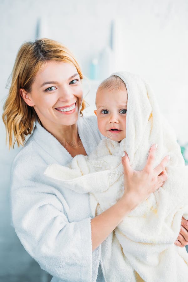 madre sonriente en la albornoz que detiene al pequeño niño confuso imagen de archivo madre sonriente en la albornoz que detiene al pequeño niño confuso imagen de archivo