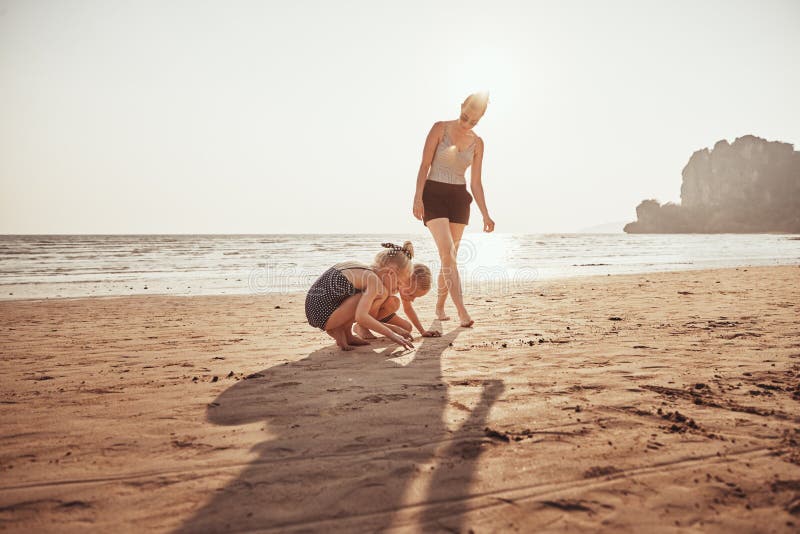 Madre sonriente que mira a sus niños el jugar en una playa arenosa fotos de archivo Madre sonriente que mira a sus niños el jugar en una playa arenosa fotos de archivo