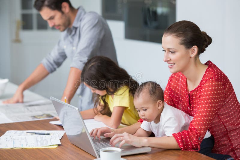 Madre sorridente che lavora al computer portatile con il bambino mentre studio della figlia immagini stock libere da diritti Madre sorridente che lavora al computer portatile con il bambino mentre studio della figlia immagini stock libere da diritti