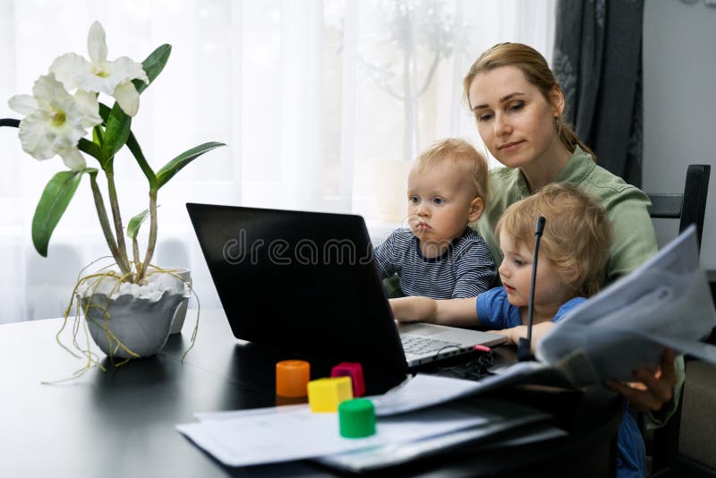 Madre trabajando desde casa usando un portátil en el escritorio con niños en su regazo imagenes de archivo