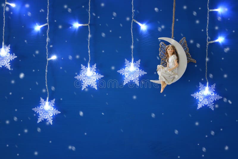 Magical christmas image of little white fairy with glitter wings sitting on the moon over blue background and silver snowflake gar