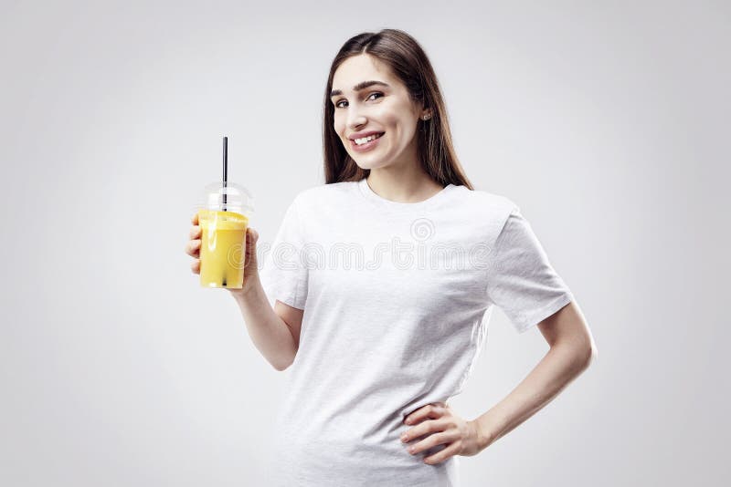 Maglietta grigia bianca d'uso della giovane bella donna che sorride con il succo d'arancia a disposizione Ritratto isolato su cen fotografia stock Maglietta grigia bianca d'uso della giovane bella donna che sorride con il succo d'arancia a disposizione Ritratto isolato su cen fotografia stock