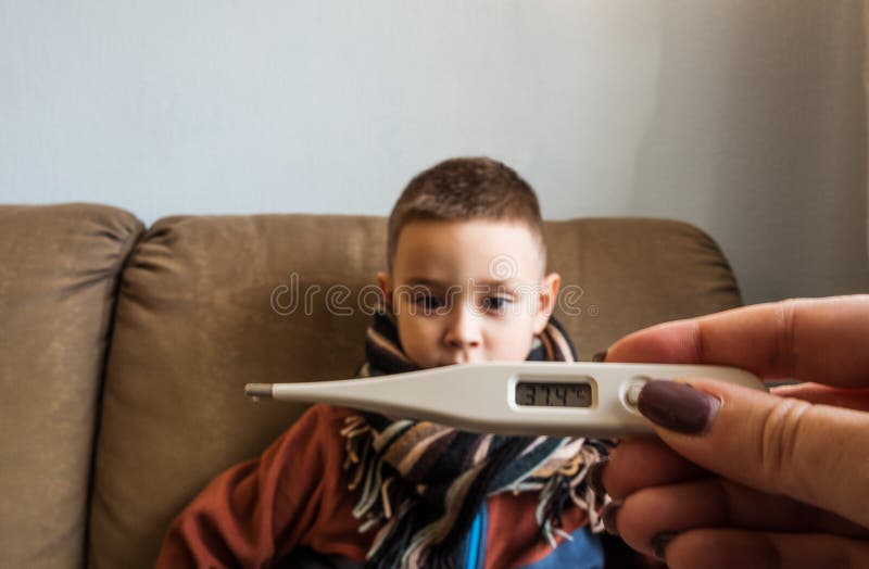 Maglione d'uso del giovane ragazzo e una sciarpa che si siede su un sofà, mentre sua madre che controlla la sua temperatura per m fotografie stock libere da diritti Maglione d'uso del giovane ragazzo e una sciarpa che si siede su un sofà, mentre sua madre che controlla la sua temperatura per m fotografie stock libere da diritti