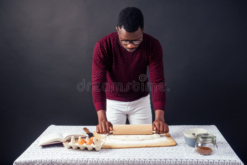 Handsome and young afro african man preparing homemade cakes American Pie from fresh dough hands dirty by flour, on the table are eggs, rolling pin and recipe book on a black background in the studio. Handsome and young afro african man preparing homemade cakes American Pie from fresh dough hands dirty by flour, on the table are eggs, rolling pin and recipe book on a black background in the studio.