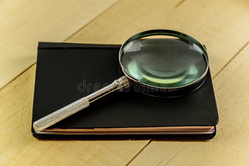 A magnifying glass and black book on top of a rustic pine table. A magnifying glass and black book on top of a rustic pine table.