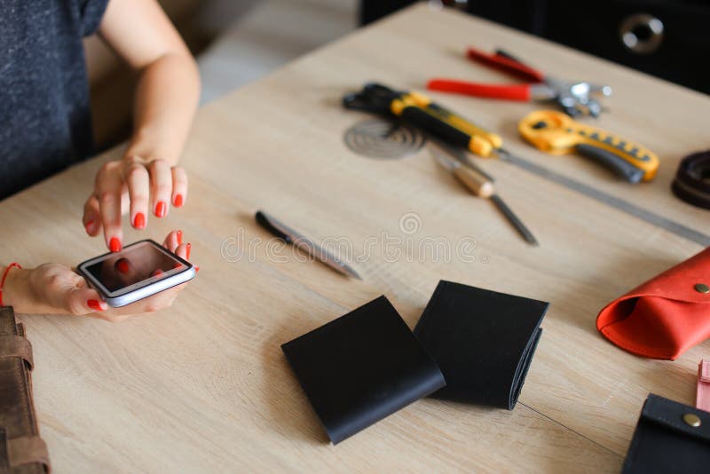 Mains femelles avec les clous rouges à l'atelier en cuir utilisant le smartphone, les portefeuilles faits main et les outils sur image stock Mains femelles avec les clous rouges à l'atelier en cuir utilisant le smartphone, les portefeuilles faits main et les outils sur image stock