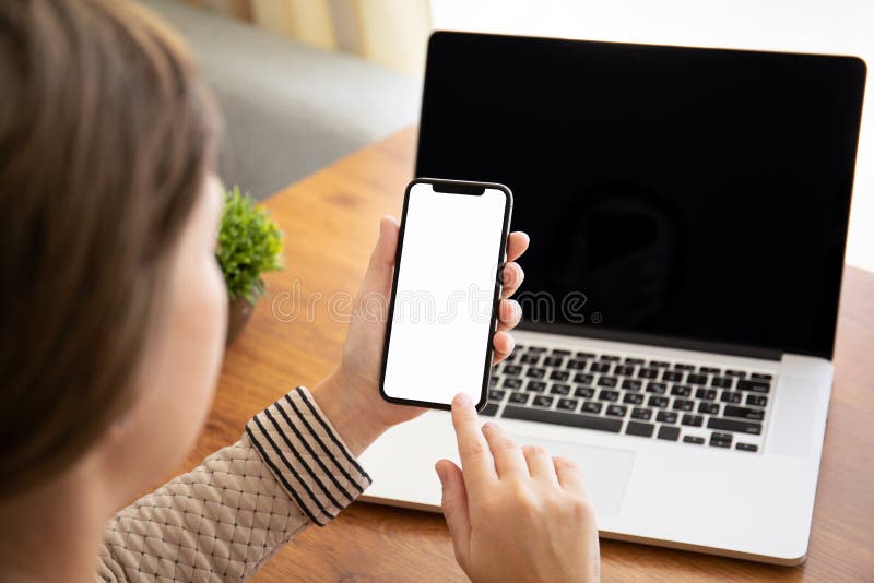 Mains femelles tenant le téléphone de contact avec l'écran d'isolement au-dessus de la table avec l'ordinateur portable dans le b photographie stock Mains femelles tenant le téléphone de contact avec l'écran d'isolement au-dessus de la table avec l'ordinateur portable dans le b photographie stock