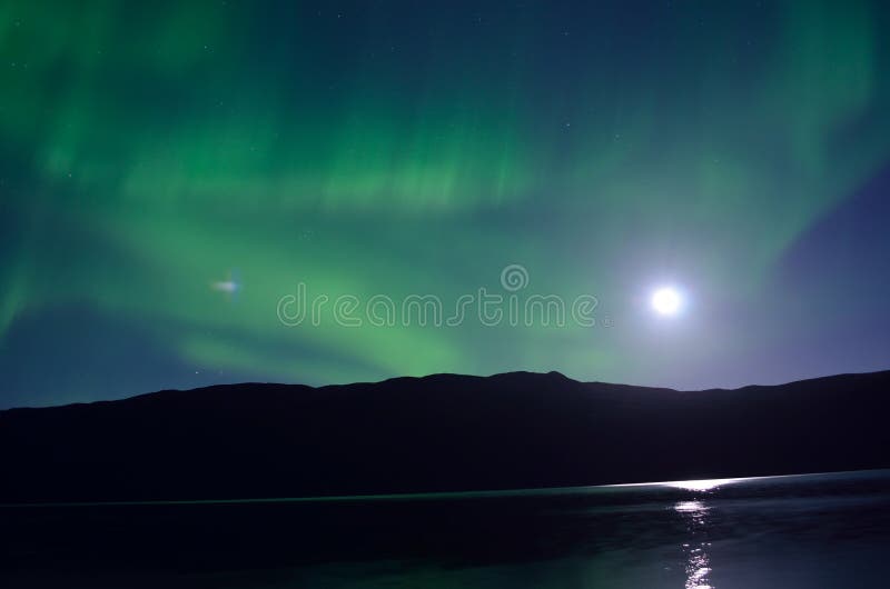 Majestic aurora borealis dancing beside full moon over mountain and calm fjord