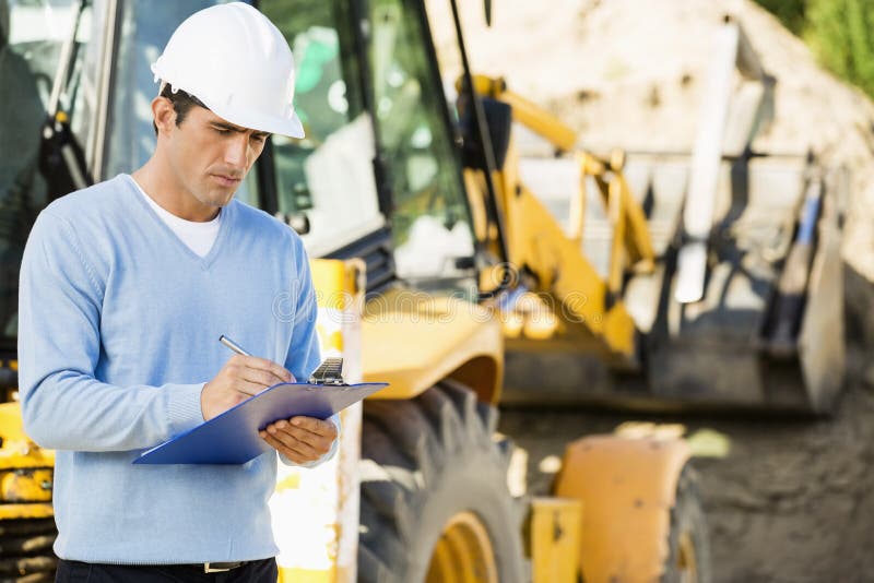 Male architect writing on clipboard against earthmover at construction site stock image