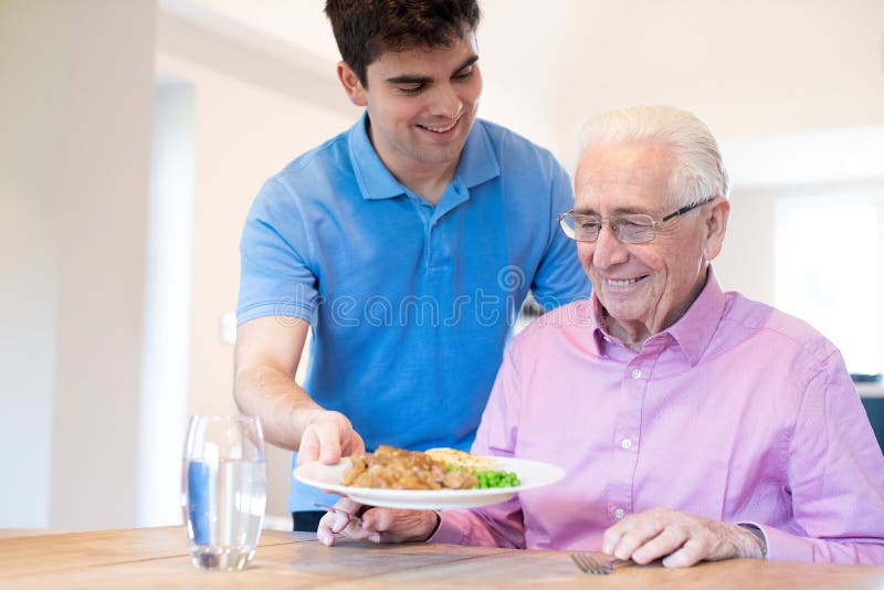 Male Care Assistant Serving Meal To Senior Male Seated At Table