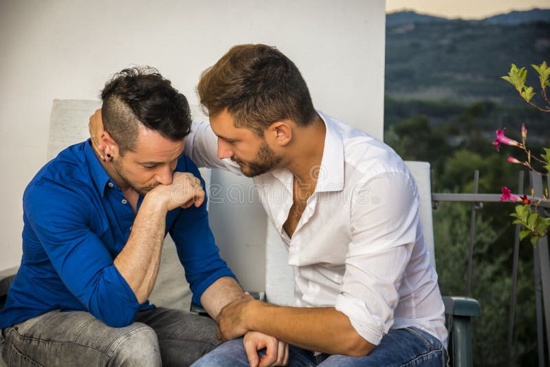 Portrait of two handsome men having trouble in relationship while sitting hugging and sad on balcony chairs. Portrait of two handsome men having trouble in relationship while sitting hugging and sad on balcony chairs.