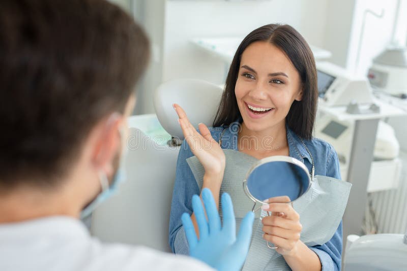 Male dentist and happy gesturing client holding mirror talking in dental office