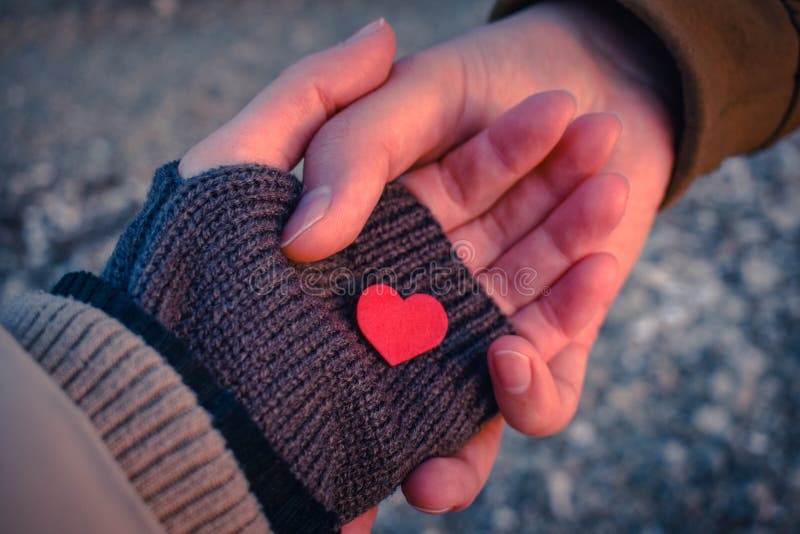 Male and female hands hold a small red heart in the sunset light.