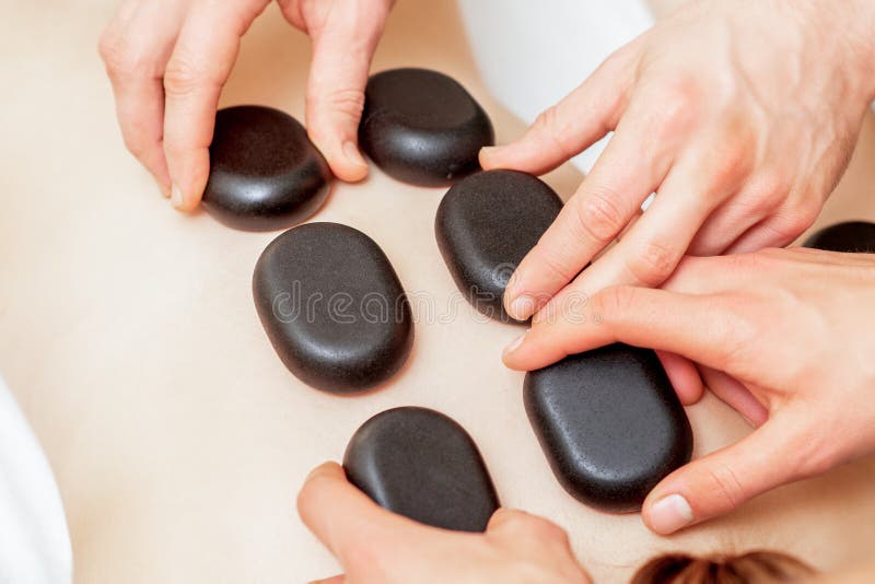 Male four hands laying stones on female back. The male four hands laying large new oval black hot stones for back massage on female back in spa salon, close up