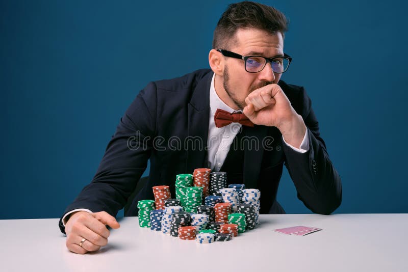 Male in glasses, black suit looking upset, sitting at white table with colored stacks of chips on it, posing on blue royalty free stock photography