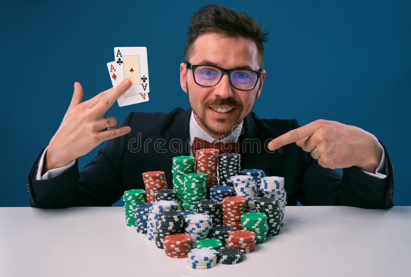 Male in glasses, black suit sitting at white table with stacks of chips, holding two playing cards, posing on blue stock photography