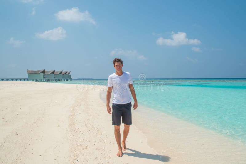 Male handsome young model in fashionable white t-shirt and black shorts at walking on sandy beach at tropical island luxury resort. In Maldives stock photography Male handsome young model in fashionable white t-shirt and black shorts at walking on sandy beach at tropical island luxury resort. In Maldives stock photography
