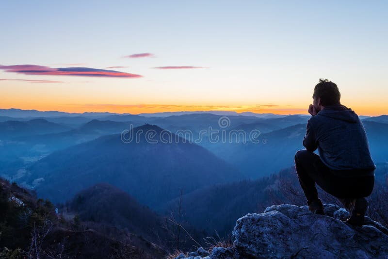 Male hiker crouching on top of the hill watching twilight landscape after sunset