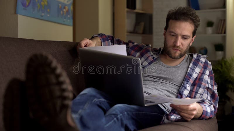 Samec ležiace na pohovka a pracovné doklady a prenosný počítač, platobné užitočnosť, fotografia fotobankového typu.