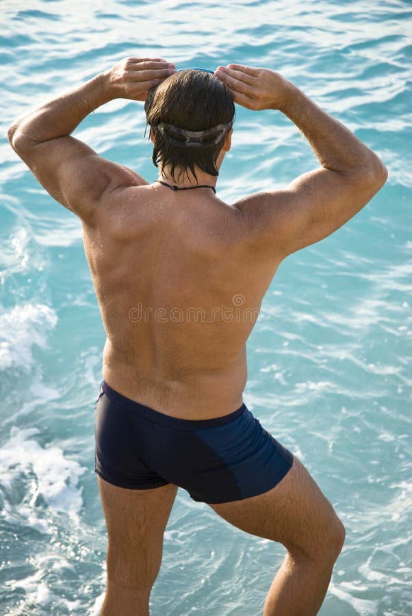 A rear view of a muscular male swimmer adjusting his swimming goggles while looking down at a beautiful turquoise sea