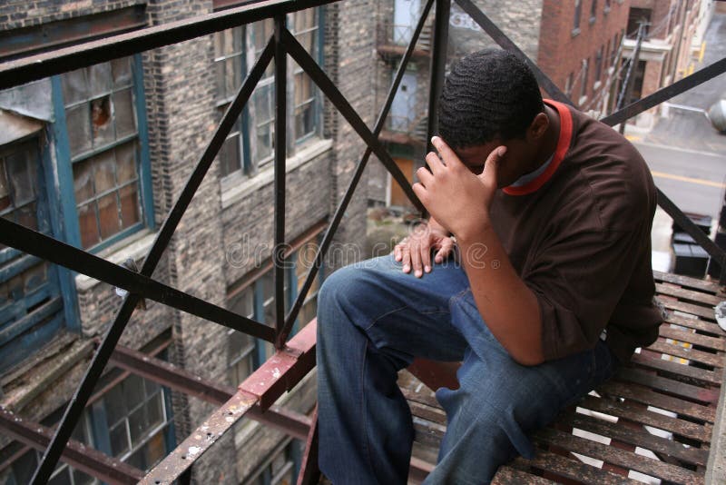 Upset African American teen male on rusty fire escape of building, head in hands. Urban setting with old buildings in background. Horizontal format. Upset African American teen male on rusty fire escape of building, head in hands. Urban setting with old buildings in background. Horizontal format.