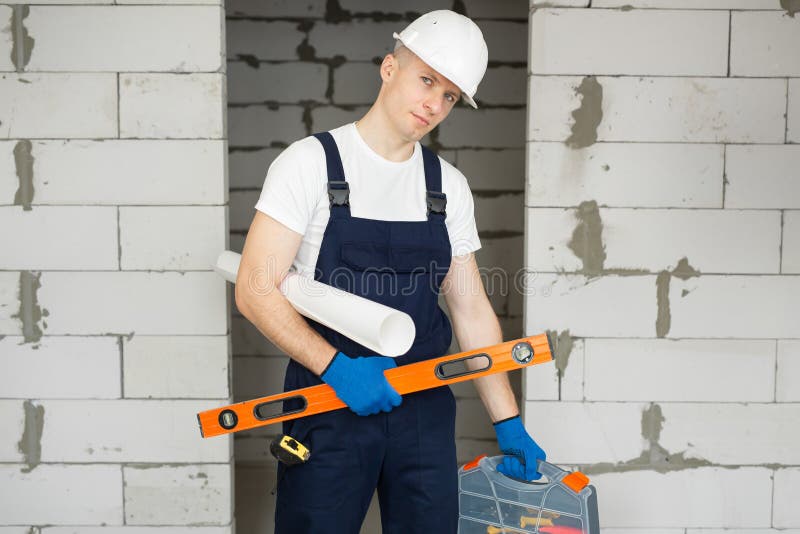 Male worker or construction worker wearing a helmet with a level, tool box, and blueprints against a brick wall.