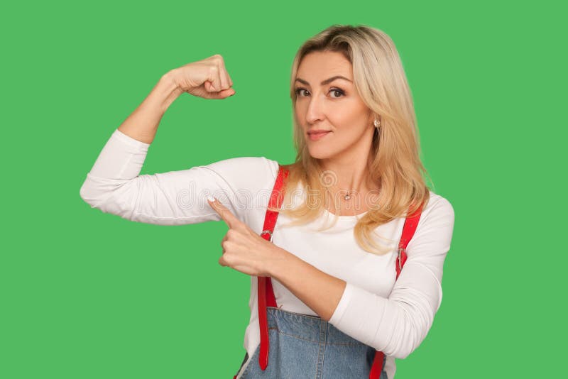 I have power! Portrait of self-confident energetic adult woman in stylish overalls raising hand pointing at biceps, feeling independent and strong. indoor studio shot isolated on green background. I have power! Portrait of self-confident energetic adult woman in stylish overalls raising hand pointing at biceps, feeling independent and strong. indoor studio shot isolated on green background