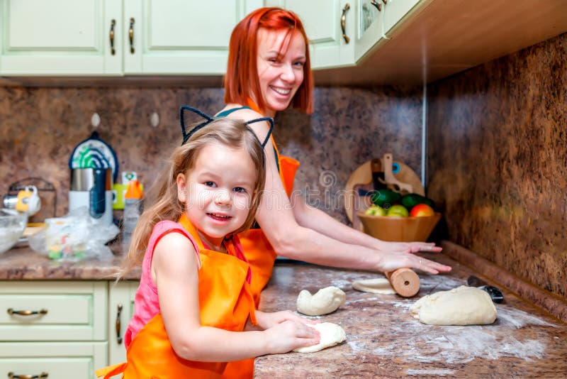 Mama i mała śliczna dziewczyna w pomarańczowych fartuchach uśmiecha się domowej roboty pizzę i robi, rolka ciasto w kuchni w domu zdjęcia stock Mama i mała śliczna dziewczyna w pomarańczowych fartuchach uśmiecha się domowej roboty pizzę i robi, rolka ciasto w kuchni w domu zdjęcia stock