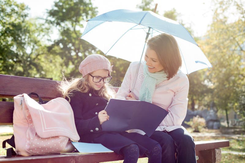 Mamma en dochter die, het lachen zitting op bank in het park, kind met het notitieboekje van de schooltaslezing spreken stock foto's Mamma en dochter die, het lachen zitting op bank in het park, kind met het notitieboekje van de schooltaslezing spreken stock foto's