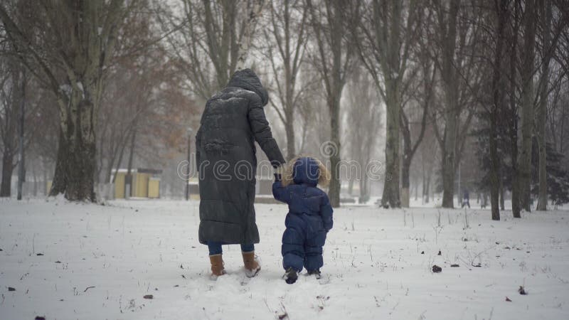 Mom and son walk through a winter park holding hands. A woman and a child walk in the snow in nature. 4k. Mom and son walk through a winter park holding hands. A woman and a child walk in the snow in nature. 4k