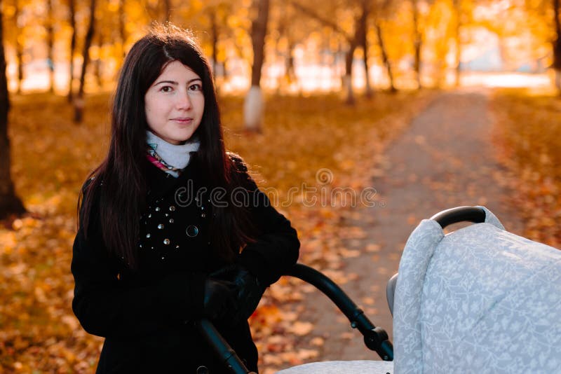 Mamá feliz joven con un cochecito en parque del otoño imagen de archivo Mamá feliz joven con un cochecito en parque del otoño imagen de archivo