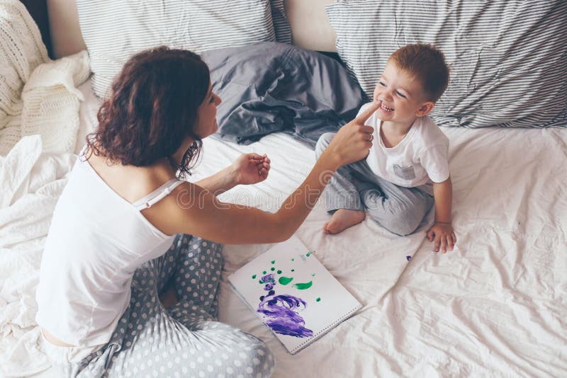 Young mother with her 2 years old little son dressed in pajamas are relaxing and playing in the bed at the weekend together, lazy morning, warm and cozy scene. Pastel colors, selective focus. Young mother with her 2 years old little son dressed in pajamas are relaxing and playing in the bed at the weekend together, lazy morning, warm and cozy scene. Pastel colors, selective focus.