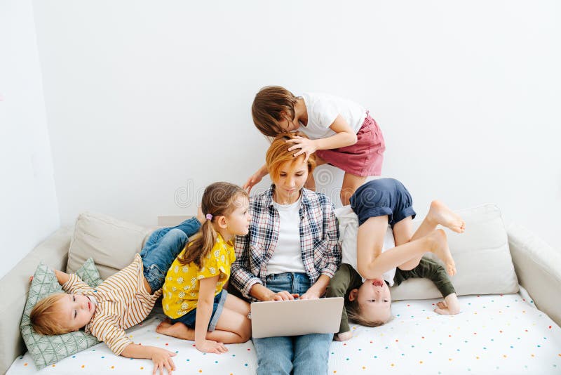 Young mom working on a laptop on a sofa in the living room, while her wild kids literally play on top of her head. They jump, roll and make noises. Young mom working on a laptop on a sofa in the living room, while her wild kids literally play on top of her head. They jump, roll and make noises