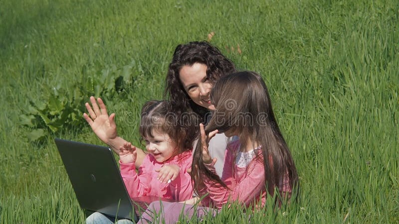Mamã com as filhas na grama verde com portátil Família em um parque em um dia ensolarado