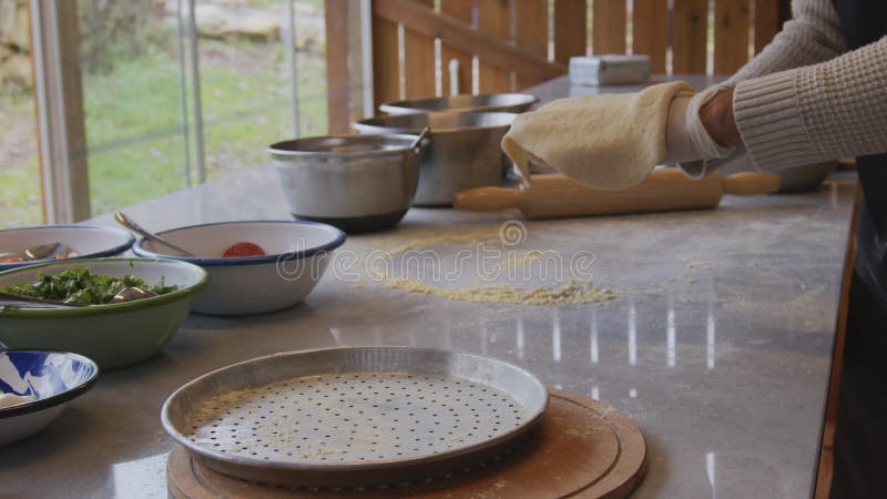 A man in an apron is rolling out pizza dough on a table sprinkled with flour. On the table in front of him are bowls of