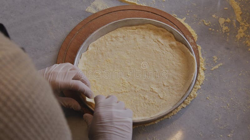 A man in an apron is rolling out pizza dough on a table sprinkled with flour. On the table in front of him are bowls of