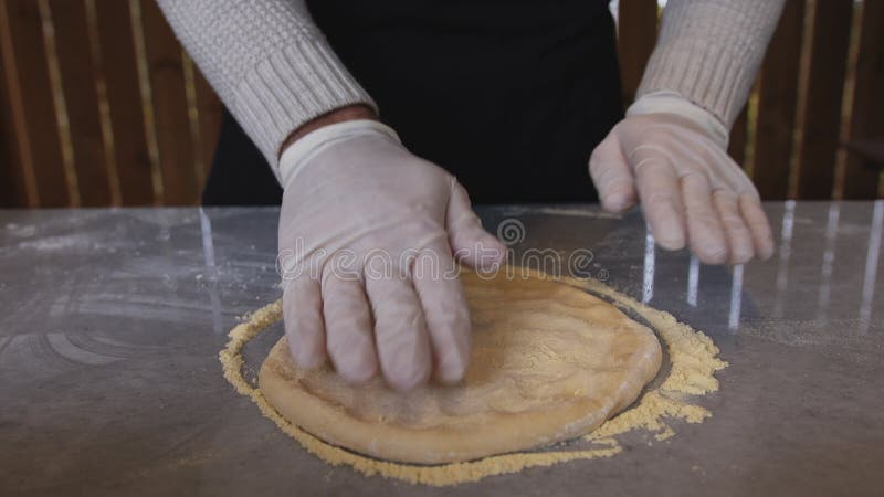 A man in an apron is rolling out pizza dough on a table sprinkled with flour. On the table in front of him are bowls of