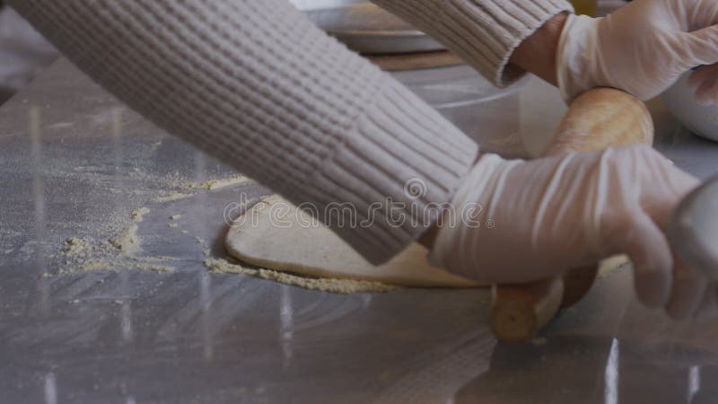 A man in an apron is rolling out pizza dough on a table sprinkled with flour. On the table in front of him are bowls of