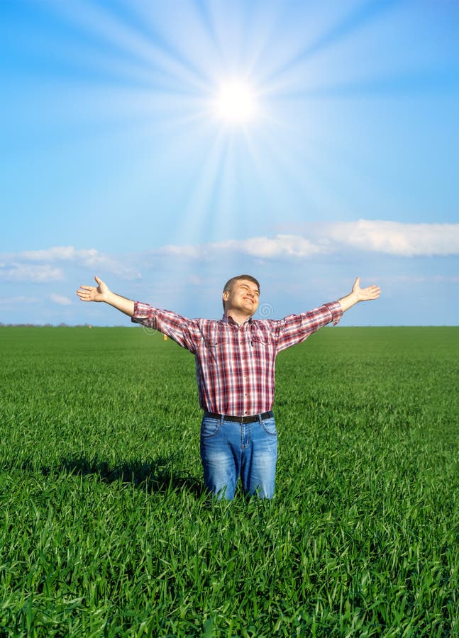 A man as a farmer poses in a field, dressed in a plaid shirt and jeans, he looks into the distance and raises his hands high in the sun