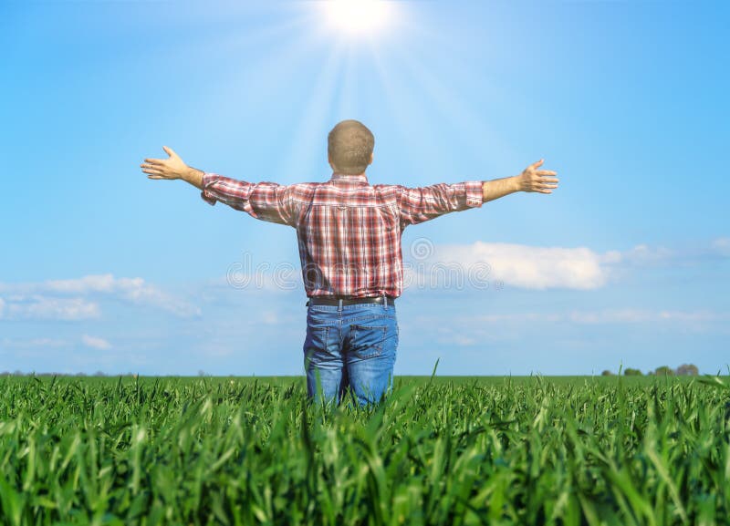 A man as a farmer poses in a field, dressed in a plaid shirt and jeans, he looks into the distance and raises his hands high in the sun