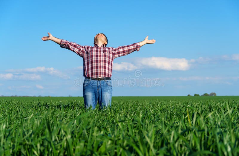 A man as a farmer poses in a field, dressed in a plaid shirt and jeans, he looks into the distance and raises his hands high in the sun