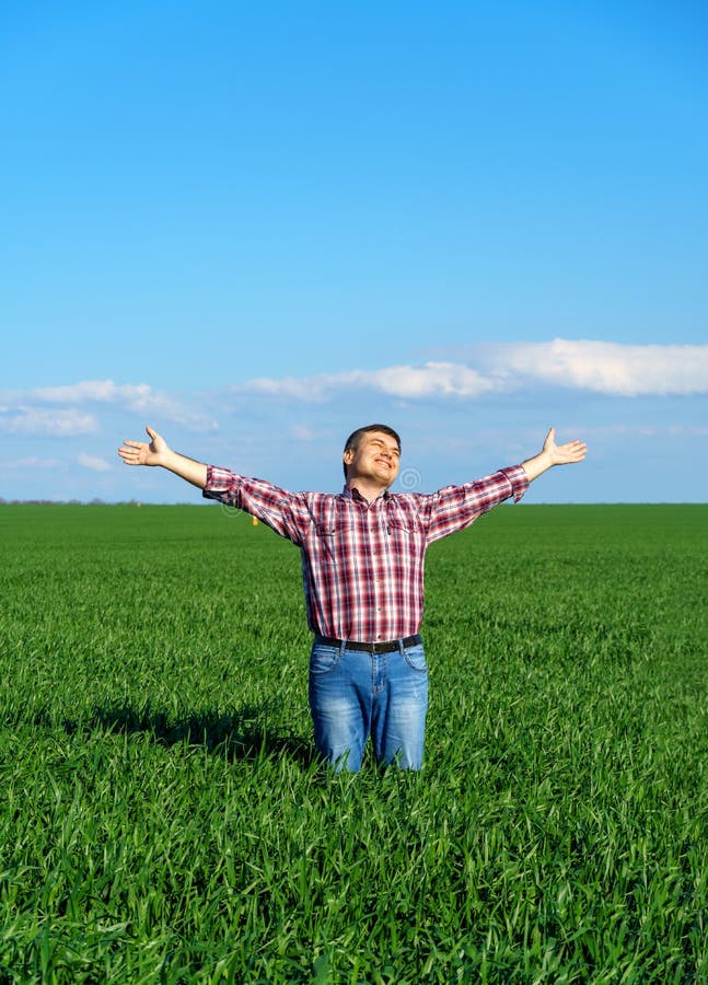 A man as a farmer poses in a field, dressed in a plaid shirt and jeans, he looks into the distance and raises his hands high in the sun