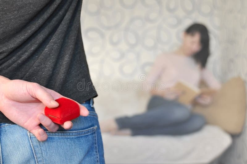 Man back and hand holding red box with marriage ring. Blurred brunette girl sitting and reading book. Engagement, valentine, propo